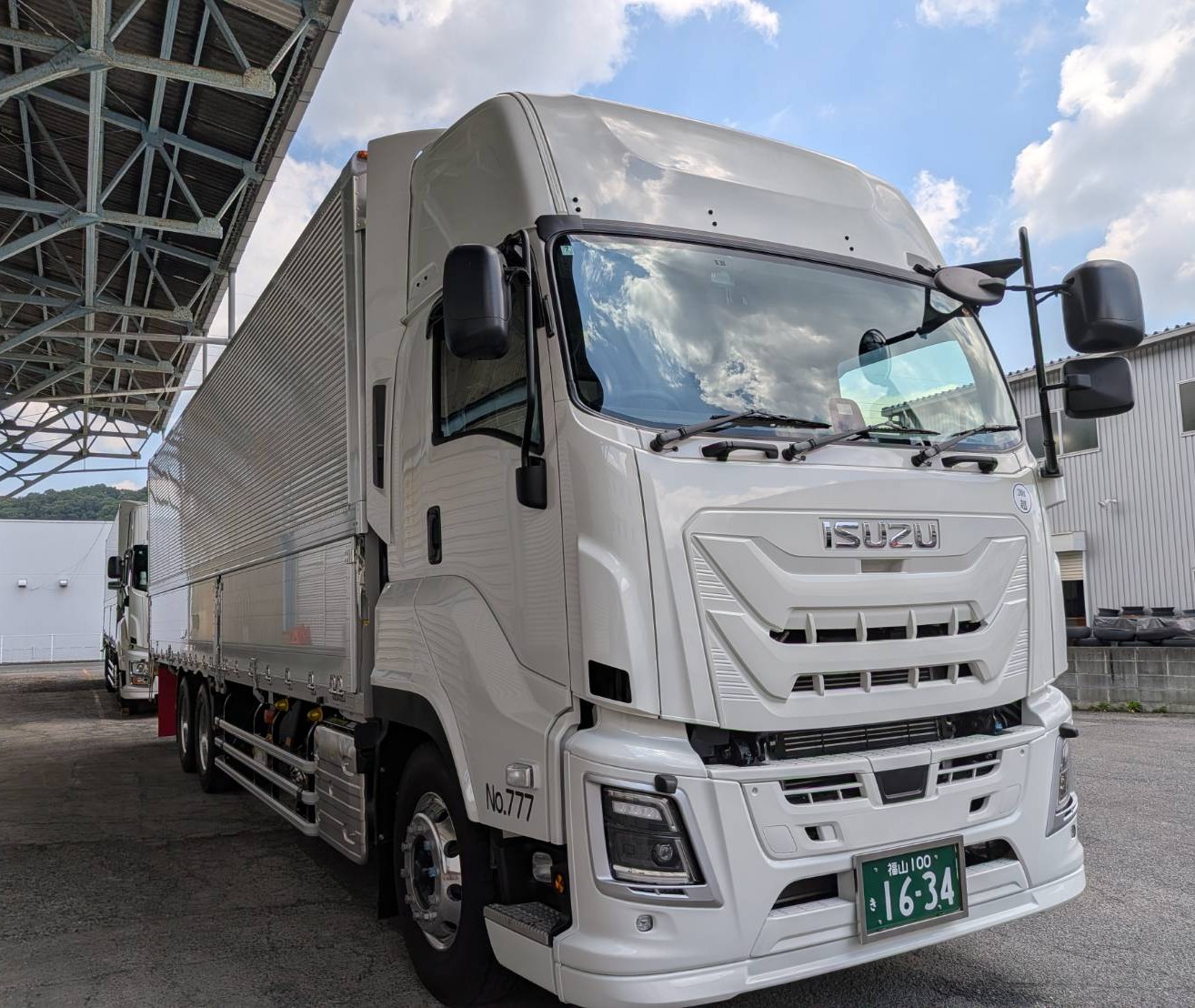 White Isuzu semi-truck with a ribbed trailer parked under a metal canopy at a loading area, front view showing grill and headlights.