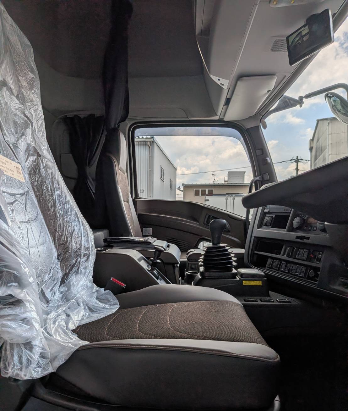 Interior of a semi-truck cab with a plastic-covered driver seat, large gear lever, and dashboard visible; exterior buildings seen through the windshield.