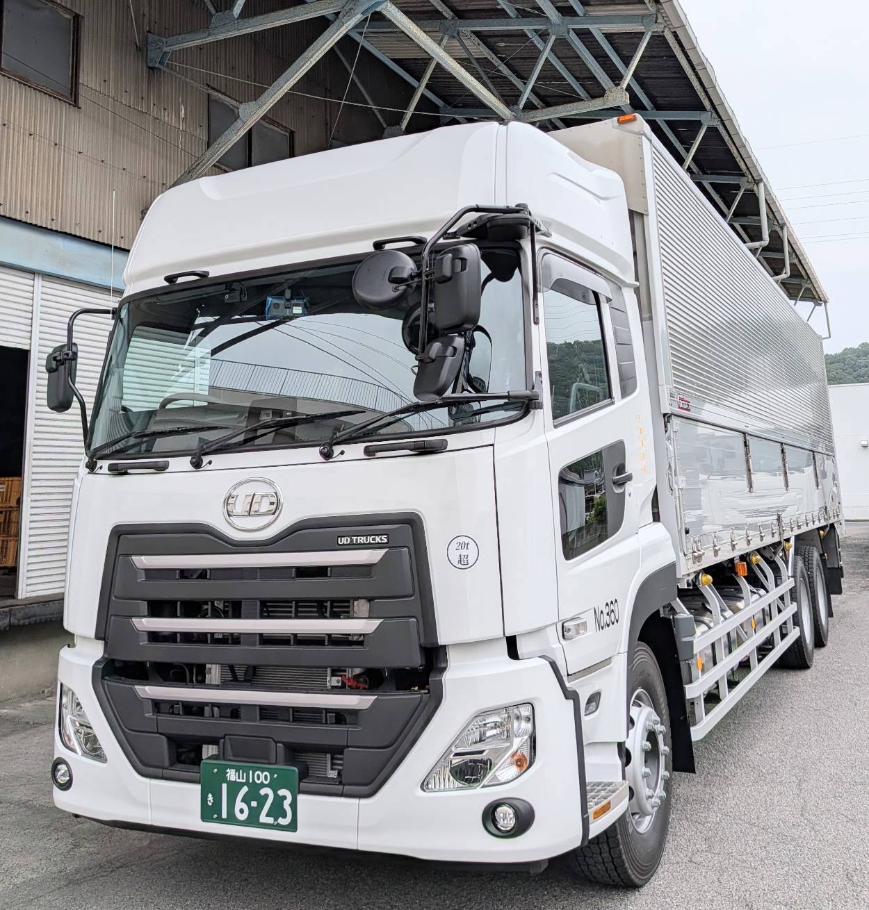 Front three-quarter view of a white UD Trucks tractor-trailer with a green license plate 16-23, parked by an industrial building.